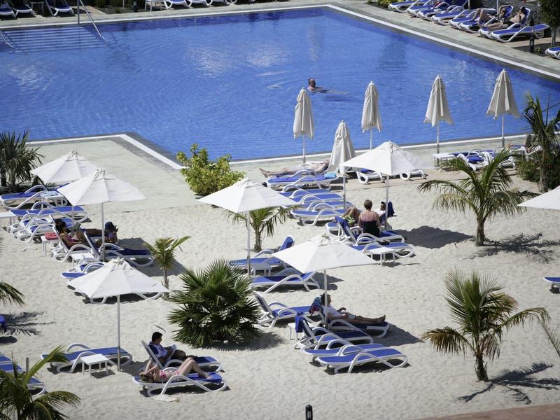 Piscina dell'hotel con lettini blu e ombrelloni bianchi circondati da piccole palme.