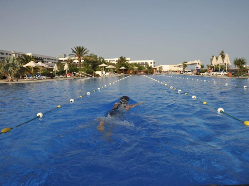 Un nuotatore in una lunga corsia di piscina all'aperto sotto un cielo azzurro con palme e edifici intorno.