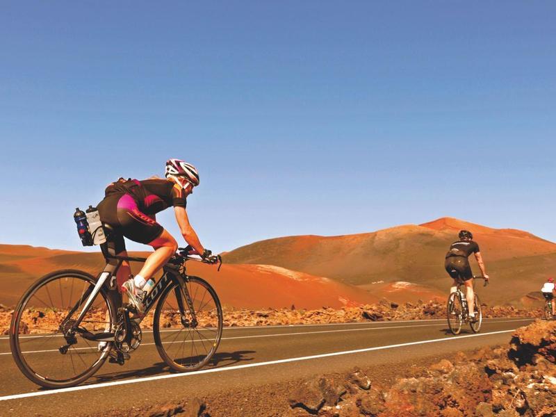 Ciclisti percorrono strade in un paesaggio desertico montuoso sotto un cielo limpido.