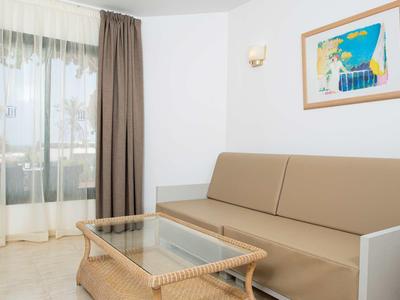 Einfache, helle Wohnzimmerecke mit braunem Sofa, Glas-Couchtisch und Fenster mit Vorhängen.