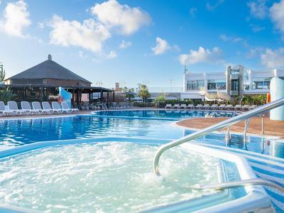 Außenwhirlpool mit sprudelndem Wasser neben großem Pool unter blauem Himmel und weißen Wolken.