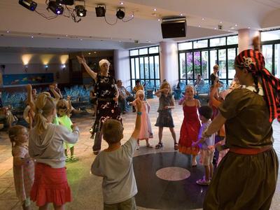 Kinder tanzen in einem hellen Raum mit großen Fenstern, begleitet von zwei Erwachsenen in Kostümen.