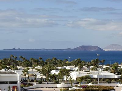 Blick auf weiße Häuser mit grünen Bäumen und blauem Meer im Hintergrund unter bewölktem Himmel.