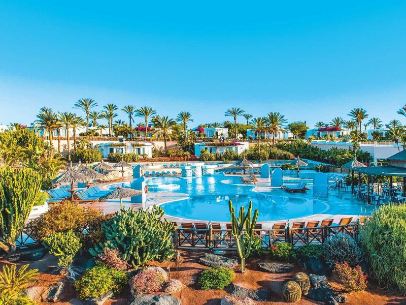 Großer Pool umgeben von Palmen und bepflanzten Beeten unter klarem blauem Himmel.