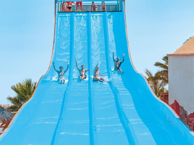 Fünf Personen rutschen nebeneinander eine breite blaue Wasserrutsche bei sonnigem Himmel hinunter.