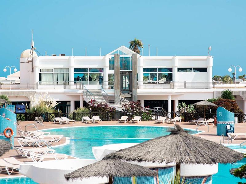 Moderner weißer mehrstöckiger Gebäudekomplex mit blauem Pool und Strohdach-Sonnenschirmen im Vordergrund.