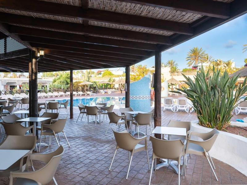 Überdachter Außenbereich mit grauen Tischen und Stühlen, im Hintergrund Pool und Palmen.