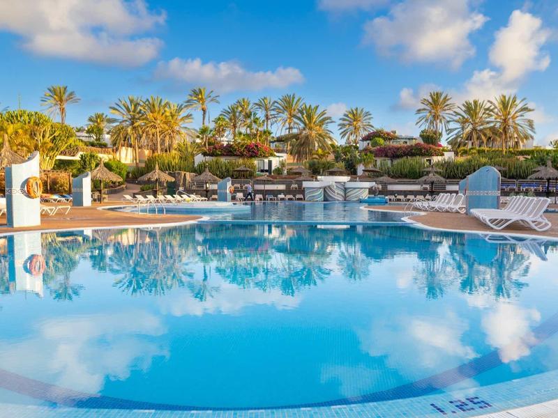Großer runder Pool mit Liegestühlen und Palmen unter blauem Himmel mit Wolken.