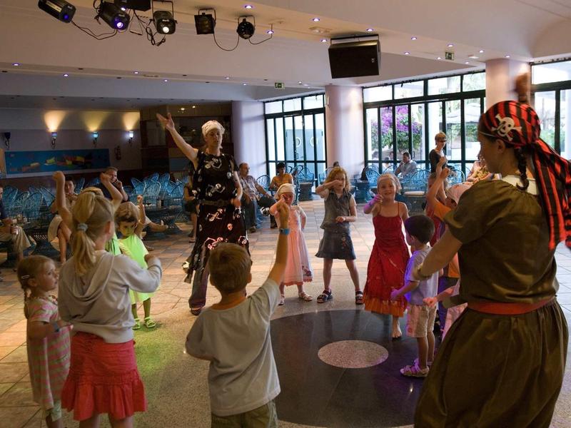 Kinder tanzen in einem hellen Raum mit großen Fenstern, begleitet von zwei Erwachsenen in Kostümen.