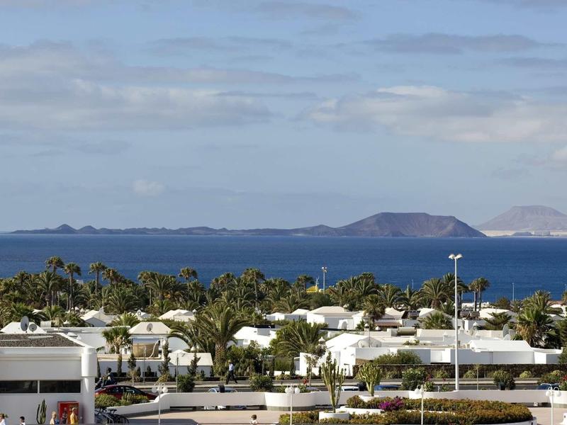 Blick auf weiße Häuser mit grünen Bäumen und blauem Meer im Hintergrund unter bewölktem Himmel.