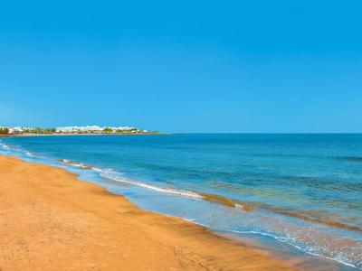 Strand mit goldenem Sand, sanften Wellen und blauem Himmel, Gebäude am Horizont sichtbar.