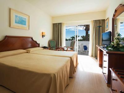 Helles Hotelzimmer mit zwei Einzelbetten, Fenster mit Balkonblick, Holzmöbeln und Stuhl.