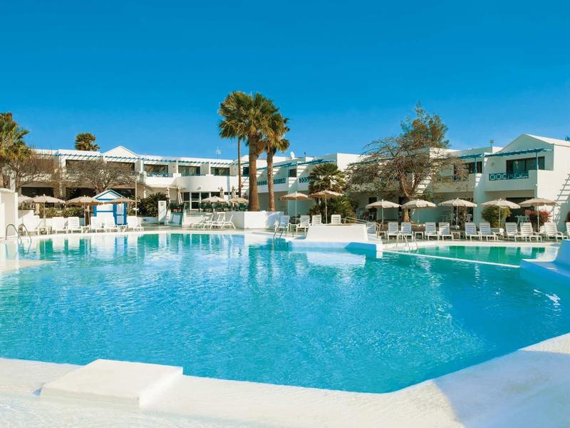 Groot hotelzwembad met ligbedden en witte gebouwen onder een heldere blauwe lucht.