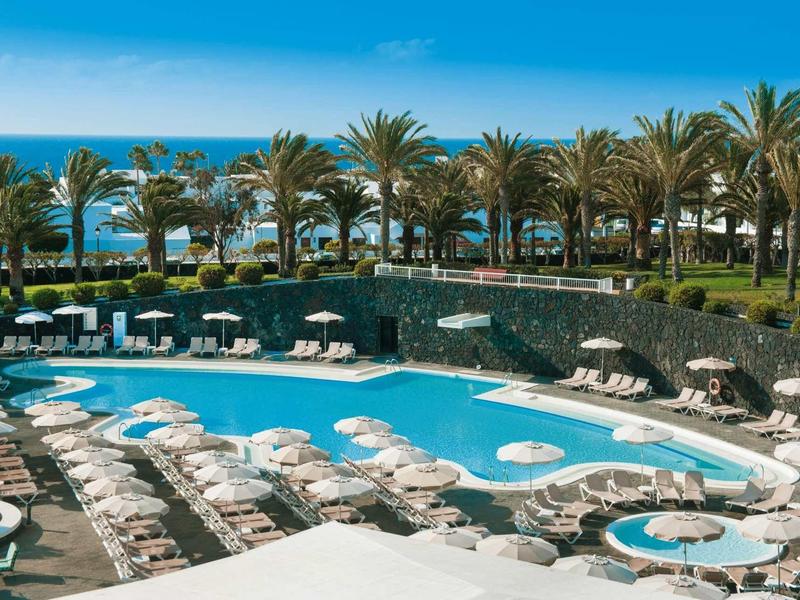 Groot hotelzwembad met ligstoelen, palmbomen en zeezicht bij heldere lucht.