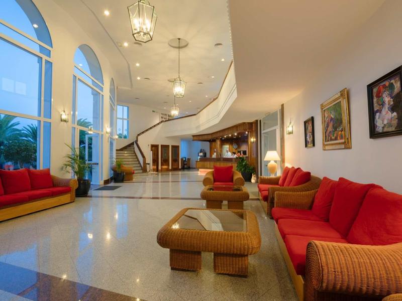 Ruime hotel-lobby met rode banken, houten tafels en hoge ramen.