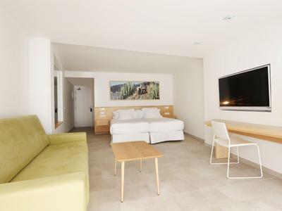 Jasny pokój hotelowy z łóżkiem podwójnym, żółtą sofą, telewizorem z płaskim ekranem i biurkiem z krzesłem.