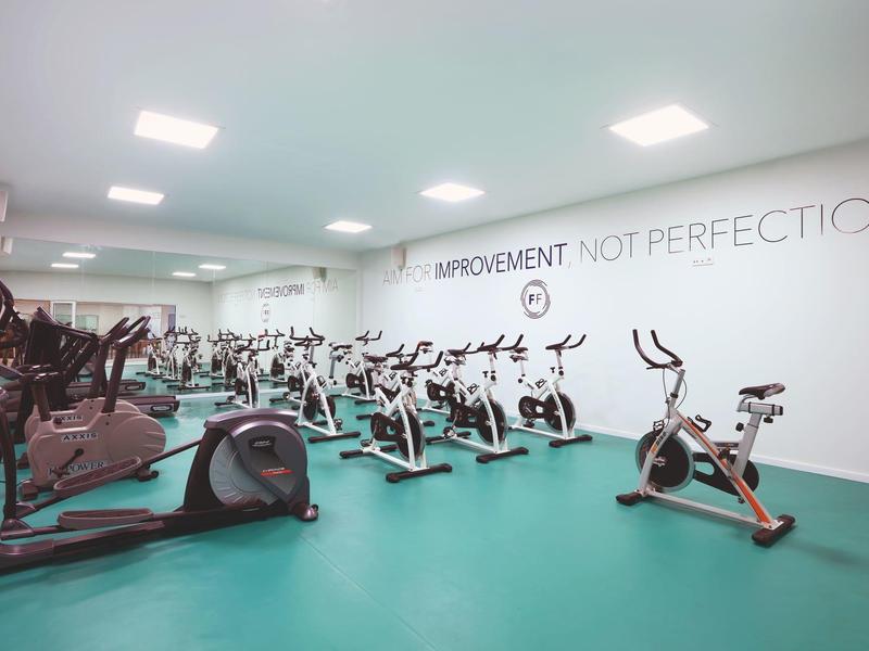 Salle de sport lumineuse avec plusieurs vélos stationnaires et tapis de course, mur blanc avec inscription.