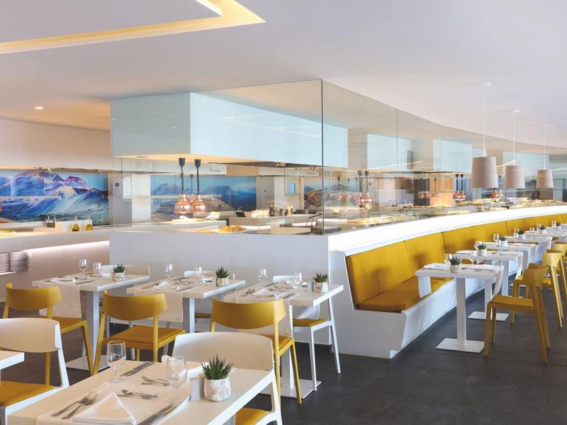 Salle à manger moderne et lumineuse avec tables blanches, chaises jaunes et buffet.