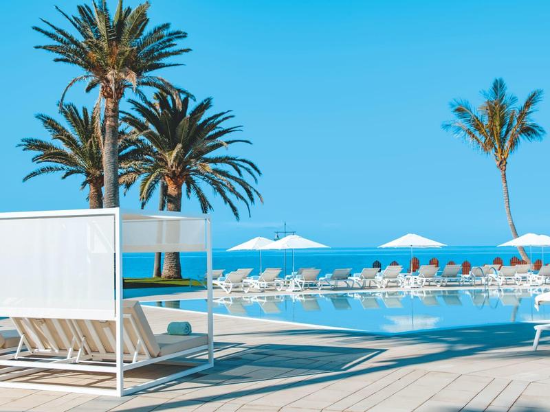 Espace piscine extérieur moderne avec chaises longues, parasols et palmiers sous un ciel clair.