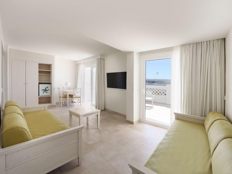 Salon lumineux avec deux canapés verts, télévision murale, grande baie vitrée et vue sur balcon.