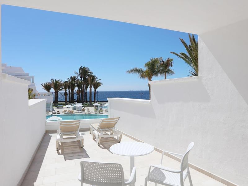 Vue depuis la terrasse avec mobilier blanc sur la piscine, les palmiers et la mer sous un ciel bleu.