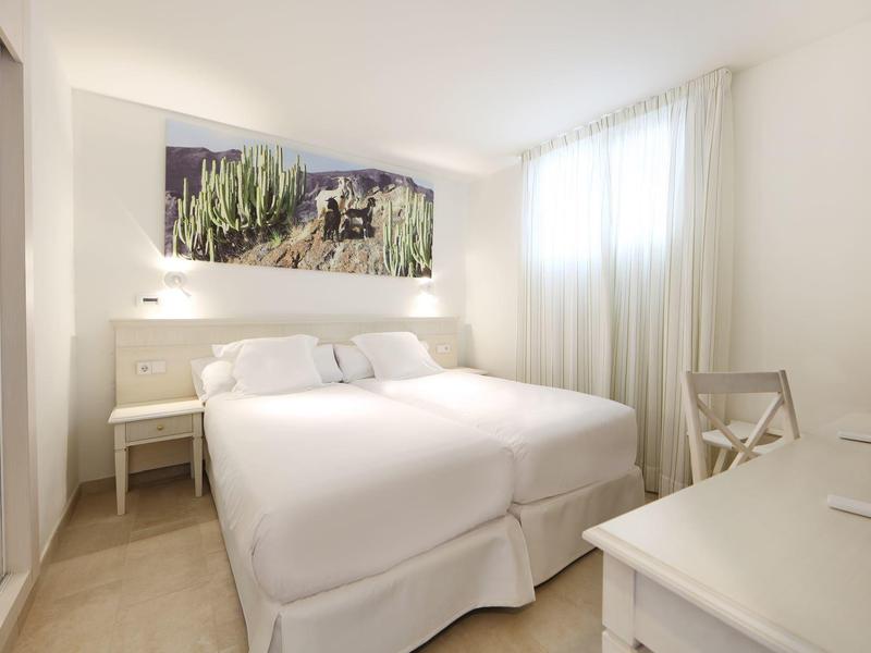 Chambre d'hôtel lumineuse avec grand lit, mobilier blanc et lumière naturelle.