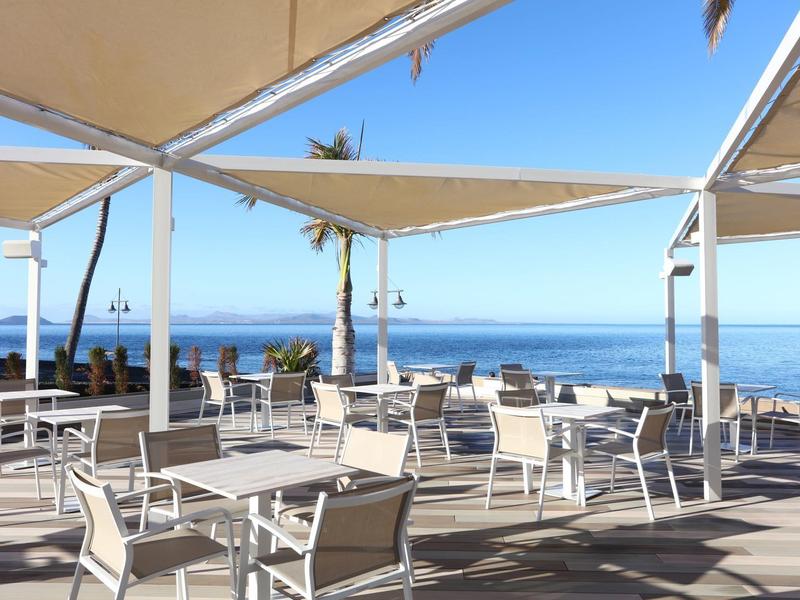 Restaurant en plein air avec tables, chaises et vue sur la mer sous des voiles d'ombrage bleues.