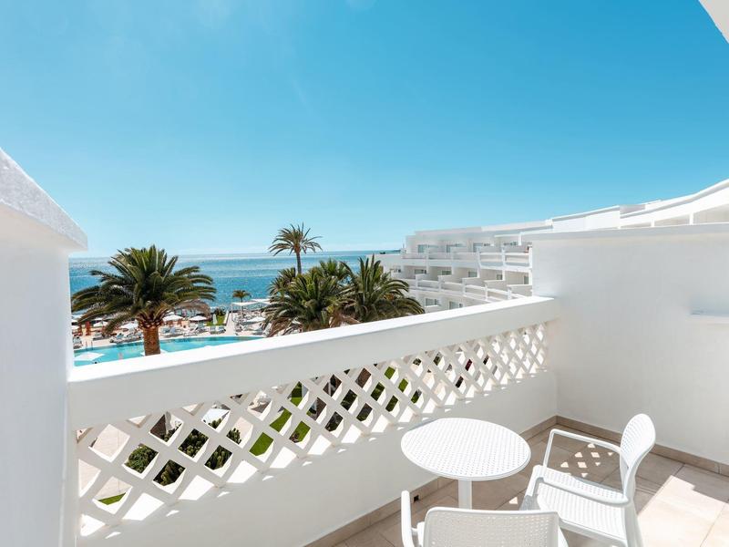 Balcon lumineux avec garde-corps blanc, chaises et vue sur des palmiers et la mer bleue.