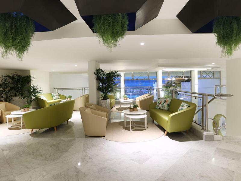 Espace lounge lumineux et moderne avec des fauteuils verts et beiges et des plantes décoratives.