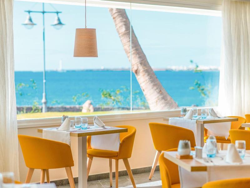 Restaurant lumineux avec des chaises jaunes et vue sur la mer par une grande fenêtre.