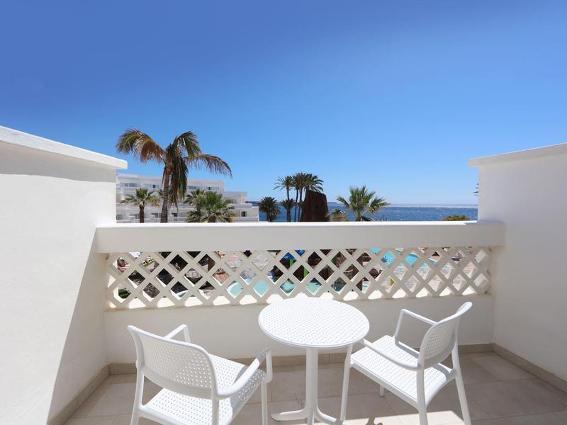 Balcon avec table et chaises blanches, vue sur des palmiers et la mer sous un ciel bleu.