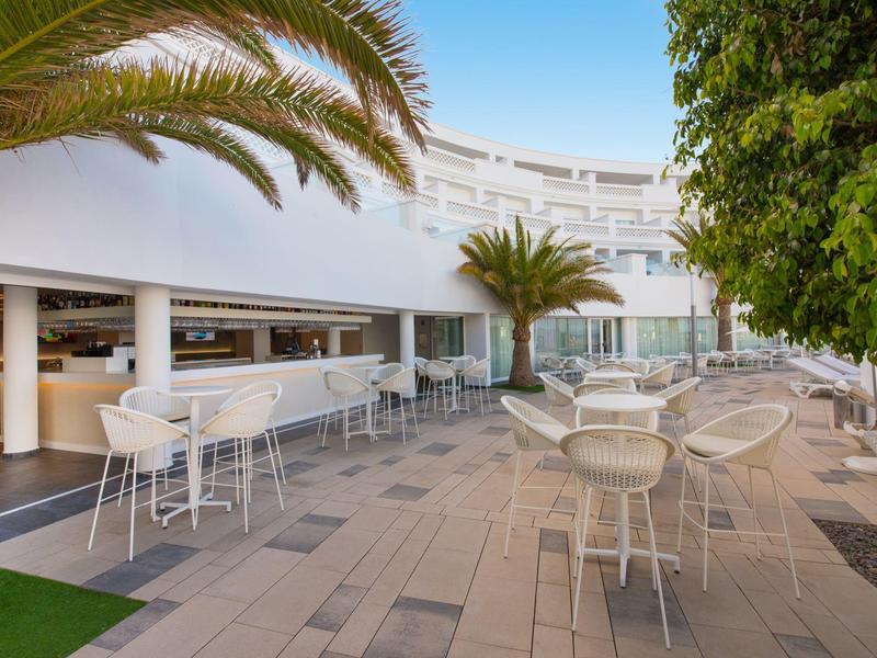Terrasse extérieure d'hôtel avec tables blanches et palmiers sous un ciel bleu clair.