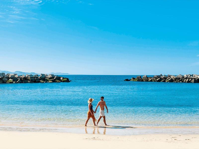 Deux personnes se tenant la main marchent sur une plage au bord d'une mer bleue claire.