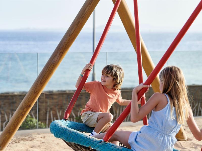 Des enfants jouent sur une balançoire nid à la plage avec la mer en arrière-plan.