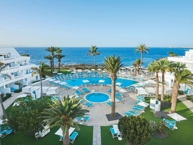 Complexe hôtelier avec grande piscine, palmiers et vue sur la mer par ciel clair.