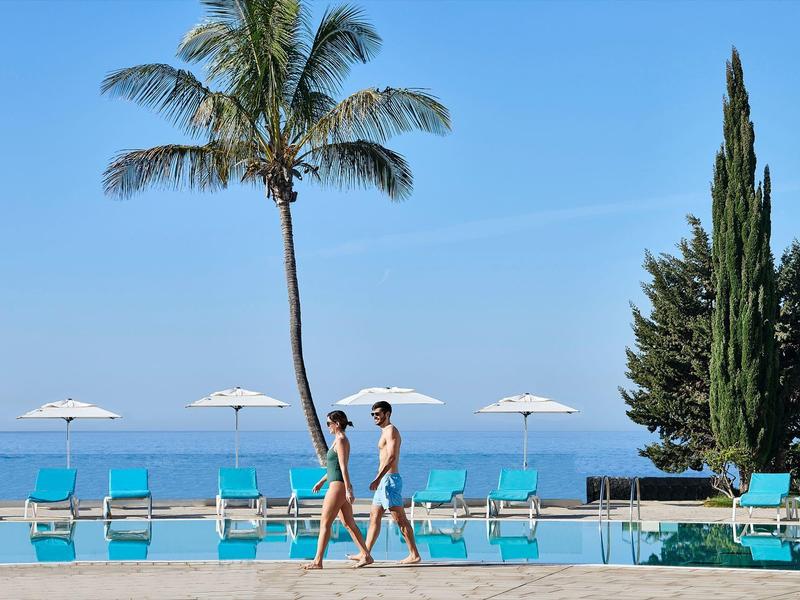 Deux personnes marchent près de la piscine avec des palmiers et parasols au bord de la mer.