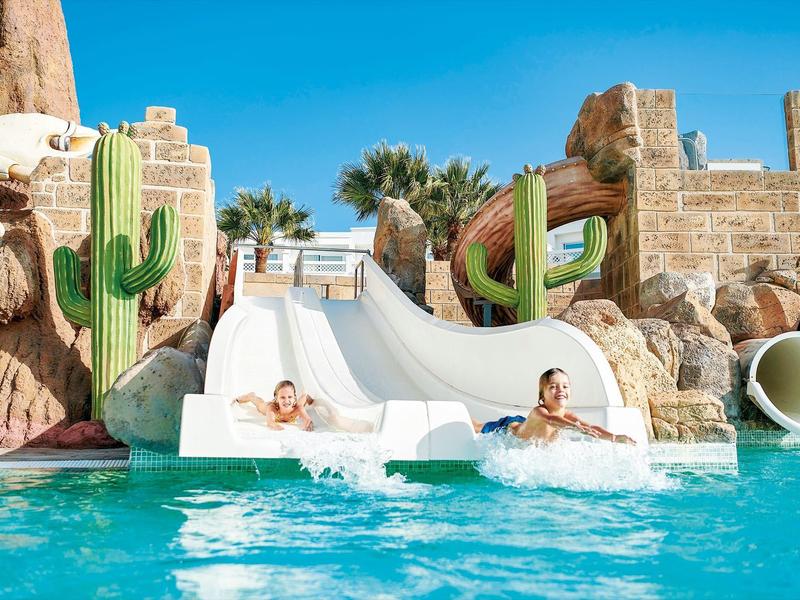Deux personnes descendent un toboggan aquatique dans une piscine décorée de cactus.