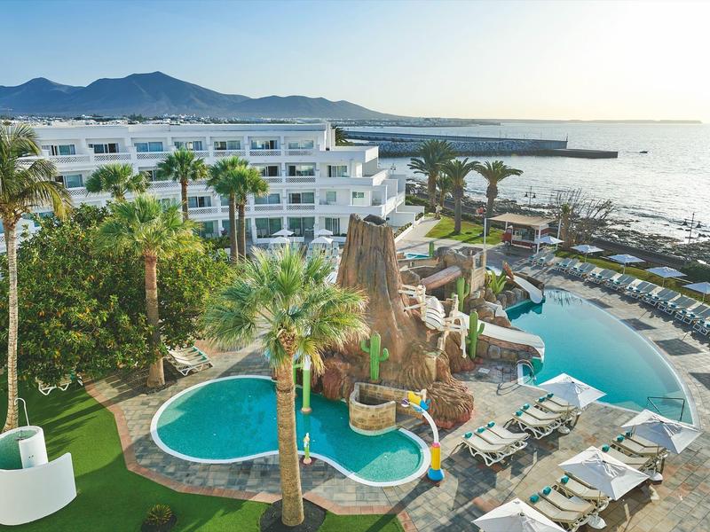 Piscine moderne d'hôtel avec palmiers, transats et vue sur la mer et les montagnes.