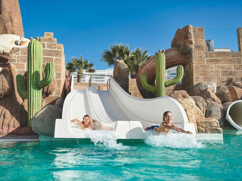 Deux personnes glissent sur un toboggan aquatique décoré de cactus dans une piscine.