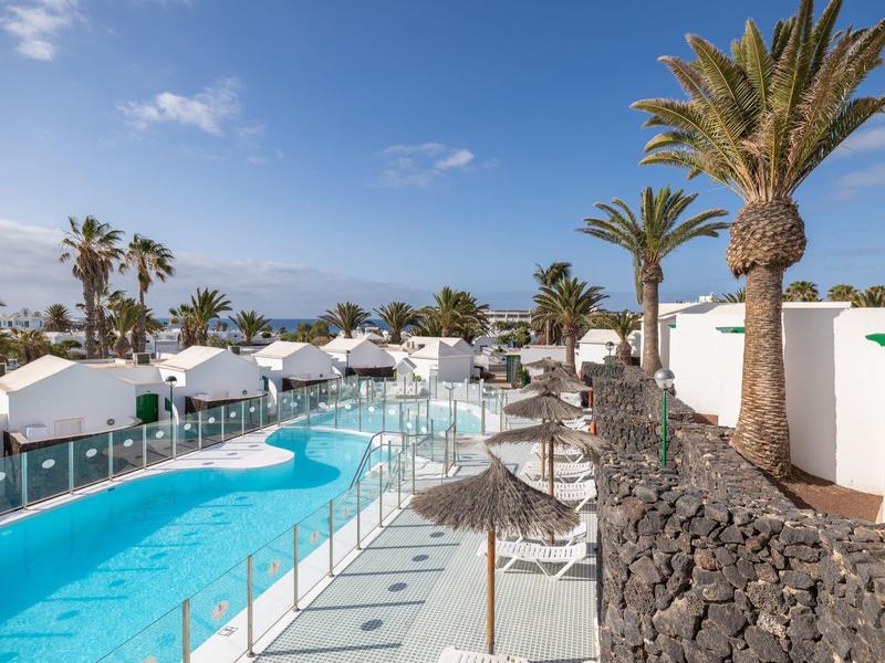 Area piscina moderna con ombrelloni e palme in un complesso alberghiero soleggiato.