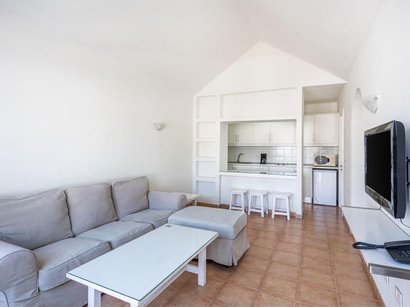 Soggiorno luminoso con divano grigio, tavolino bianco, TV e cucina a vista con sgabelli.