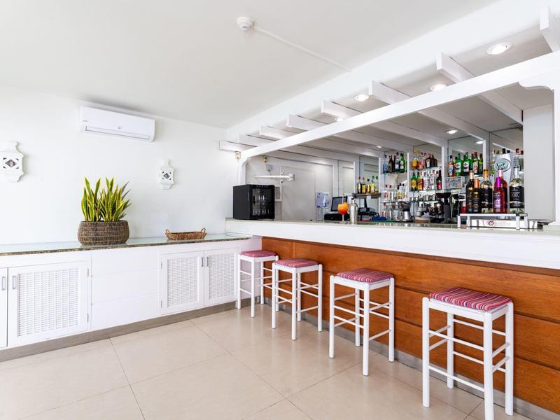 Zona bar luminosa con banco in legno, sgabelli bianchi e piante su mobile bianco.