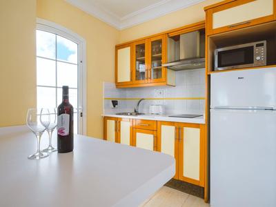 Mała kuchnia z białymi i drewnianymi szafkami, lodówka, mikrofalówka oraz stół z butelką wina i kieliszkami.