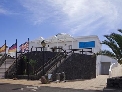 Modernes weißes Gebäude mit schwarzer Treppe, mehreren Flaggen und Palmen bei blauem Himmel.