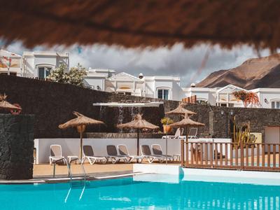 Bazénová oblast s lehátky a slunečníky před bílými budovami a horami v pozadí.