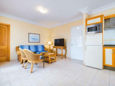 Jasny salon z niebieskimi fotelami, telewizorem i aneksem kuchennym w apartamencie wakacyjnym.