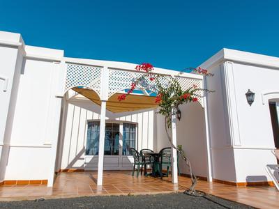 Biały domek wakacyjny z tarasem, miejscem do siedzenia i kwitnącym drzewem pod jasnym niebieskim niebem.