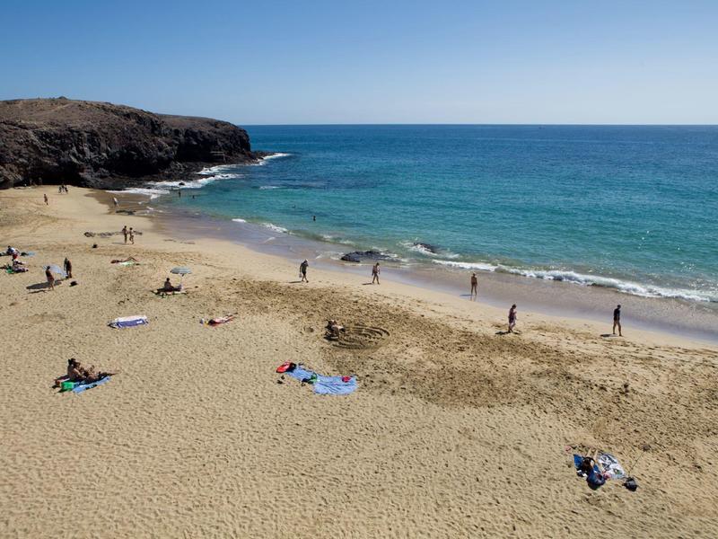 Piaszczysta plaża z kilkoma osobami, skaliste wybrzeże i spokojne niebieskie morze pod czystym niebem.