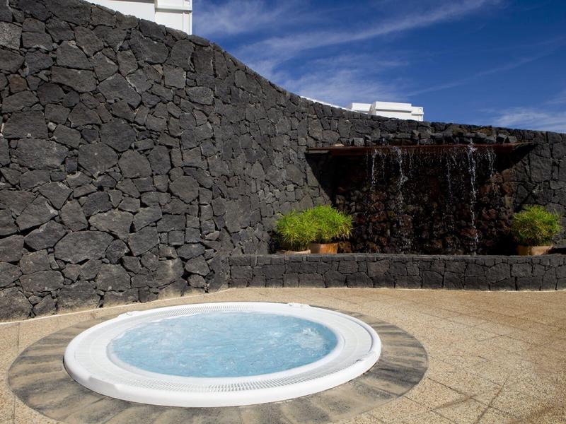 Jacuzzi z wodospadem przed czarną kamienną ścianą pod błękitnym niebem.