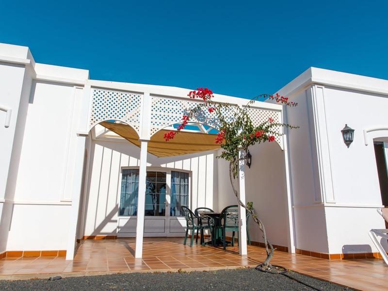 Biały domek wakacyjny z tarasem, miejscem do siedzenia i kwitnącym drzewem pod jasnym niebieskim niebem.